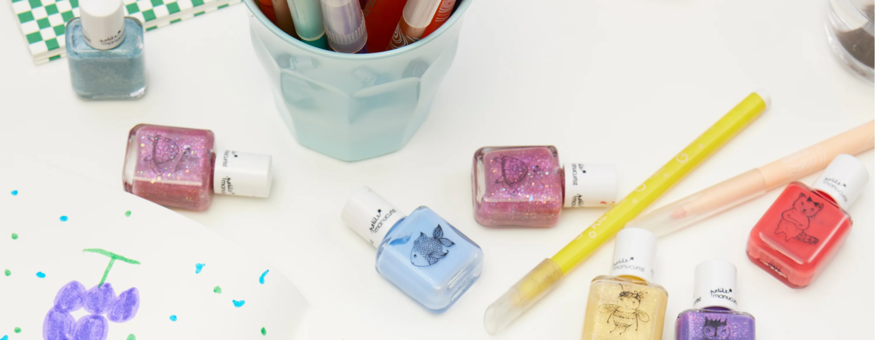 Several colorful bottles of nail polish with animal illustrations, two pens, a cup of markers, and a child’s drawing are arranged on a white surface.
