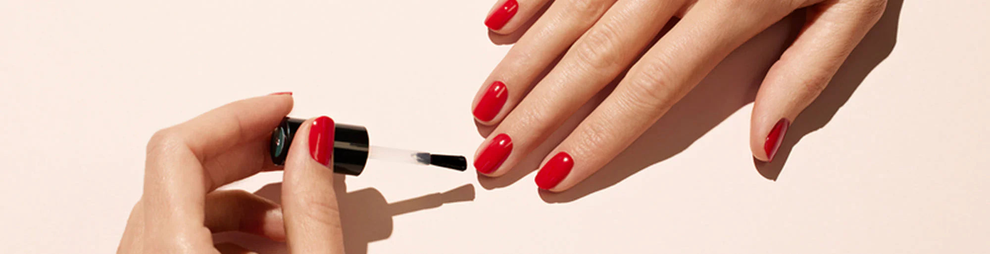 A hand with red nail polish is being painted with a clear top coat using a nail polish brush on a light background.