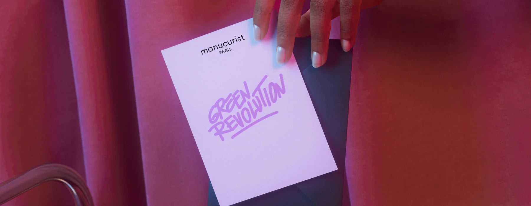A hand with pastel-colored nails holds a card reading manucurist paris and GREEN REVOLUTION in purple against a pink and red background.