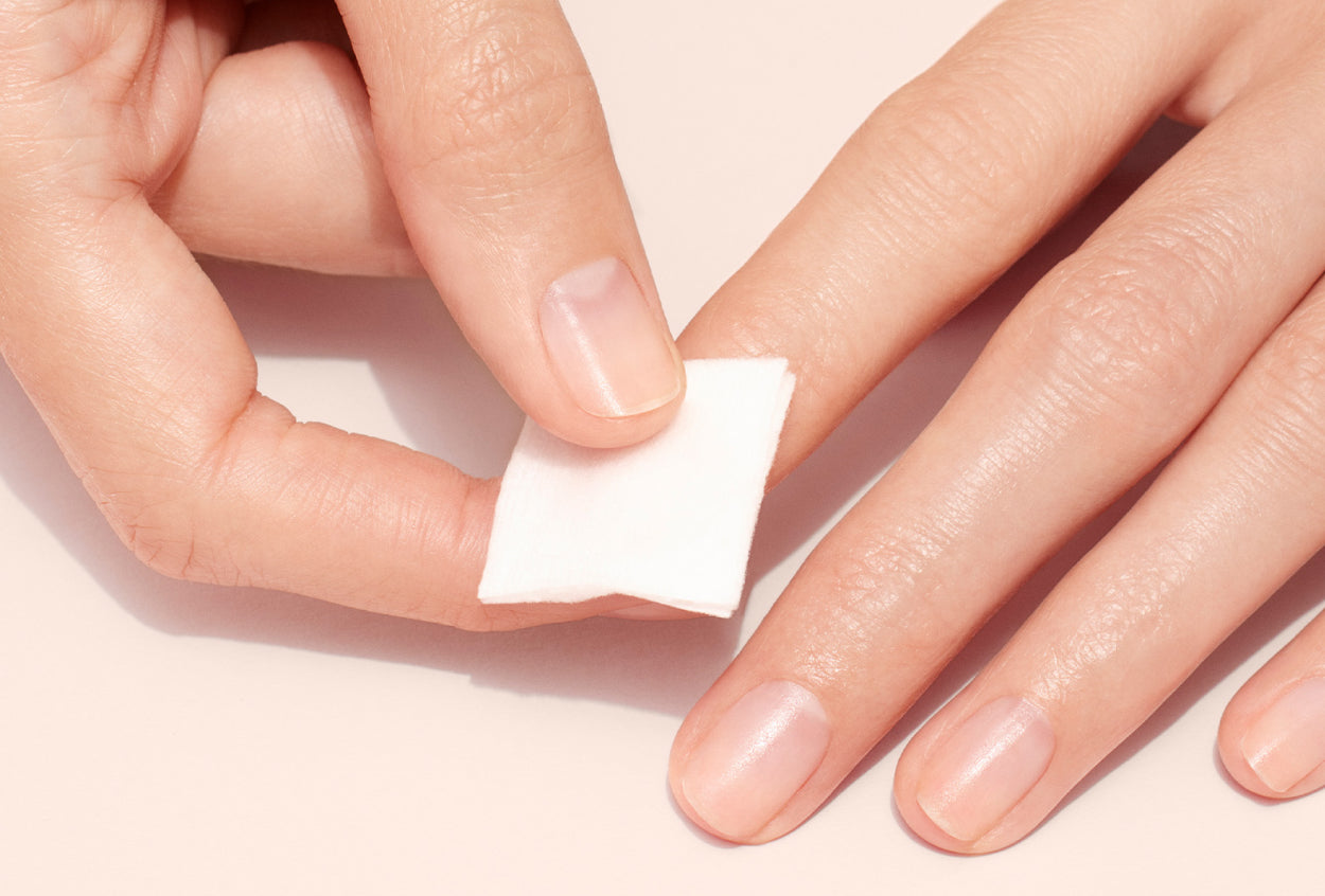 A hand holds a white cotton pad, gently cleaning or wiping the nails of the other hand, which has clean, natural fingernails on a light background.