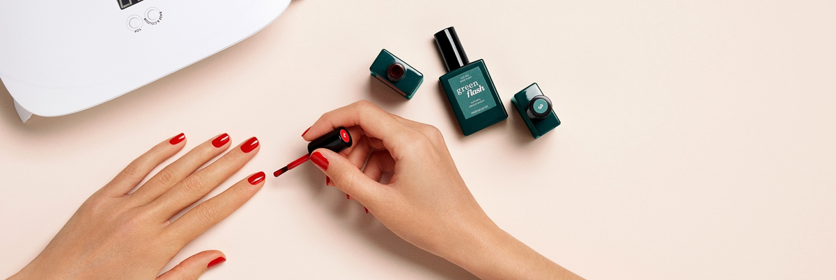 A person is painting their fingernails with red nail polish. Three bottles of nail polish and a white UV nail lamp are on the light-colored table.