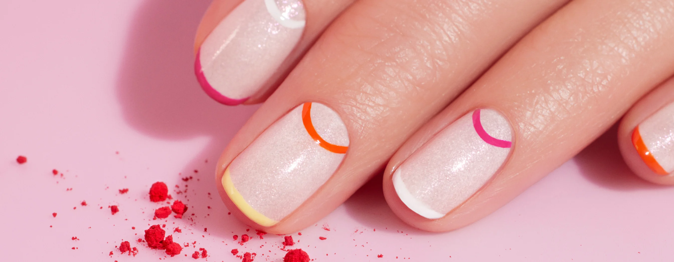 A hand with shimmery nude nail polish features colorful, thin curved lines at the tips of each nail. Red powder is scattered on the pink background.