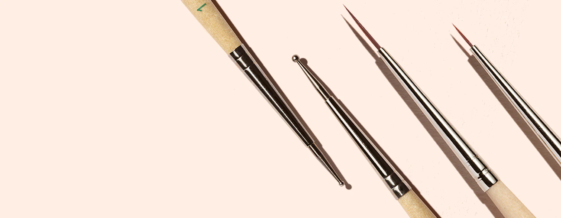 Four nail art brushes with wooden handles and metal tips are arranged diagonally on a light beige background, casting soft shadows.