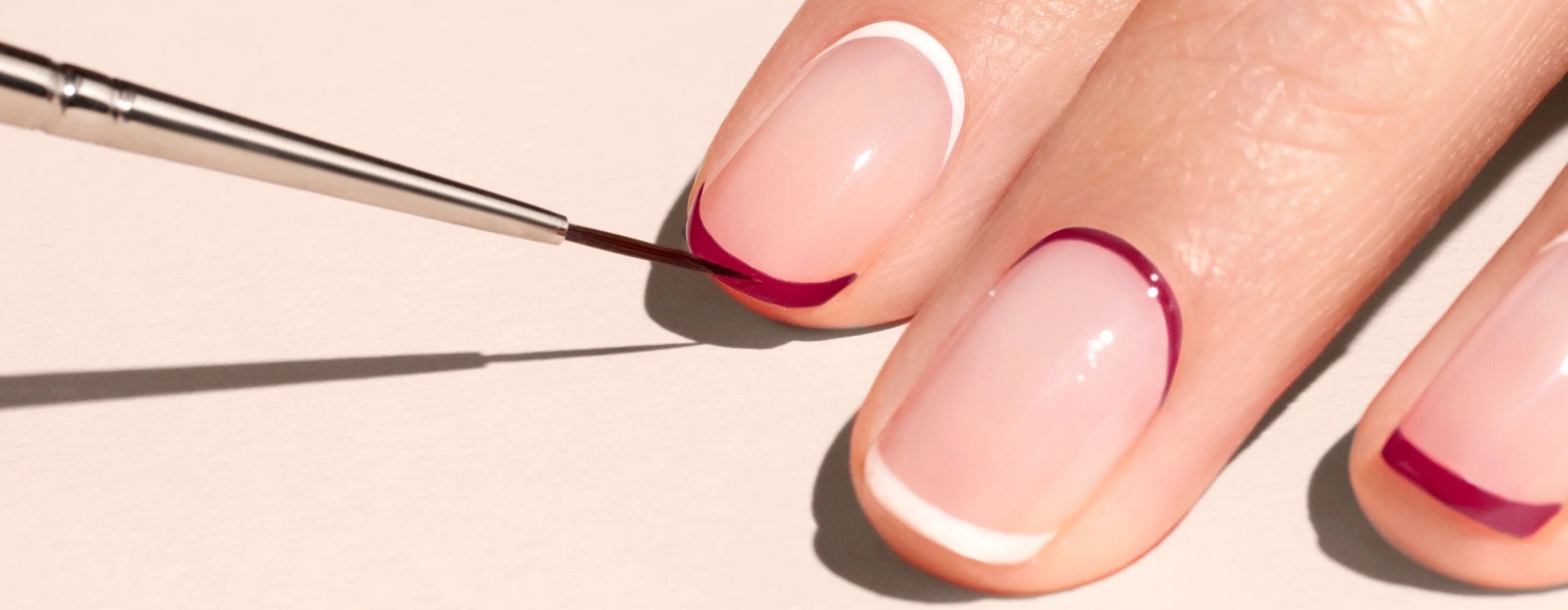 Close-up of a hand with neatly manicured nails, featuring a white and burgundy French tip design. A thin brush is being used to paint burgundy polish on one nail tip.