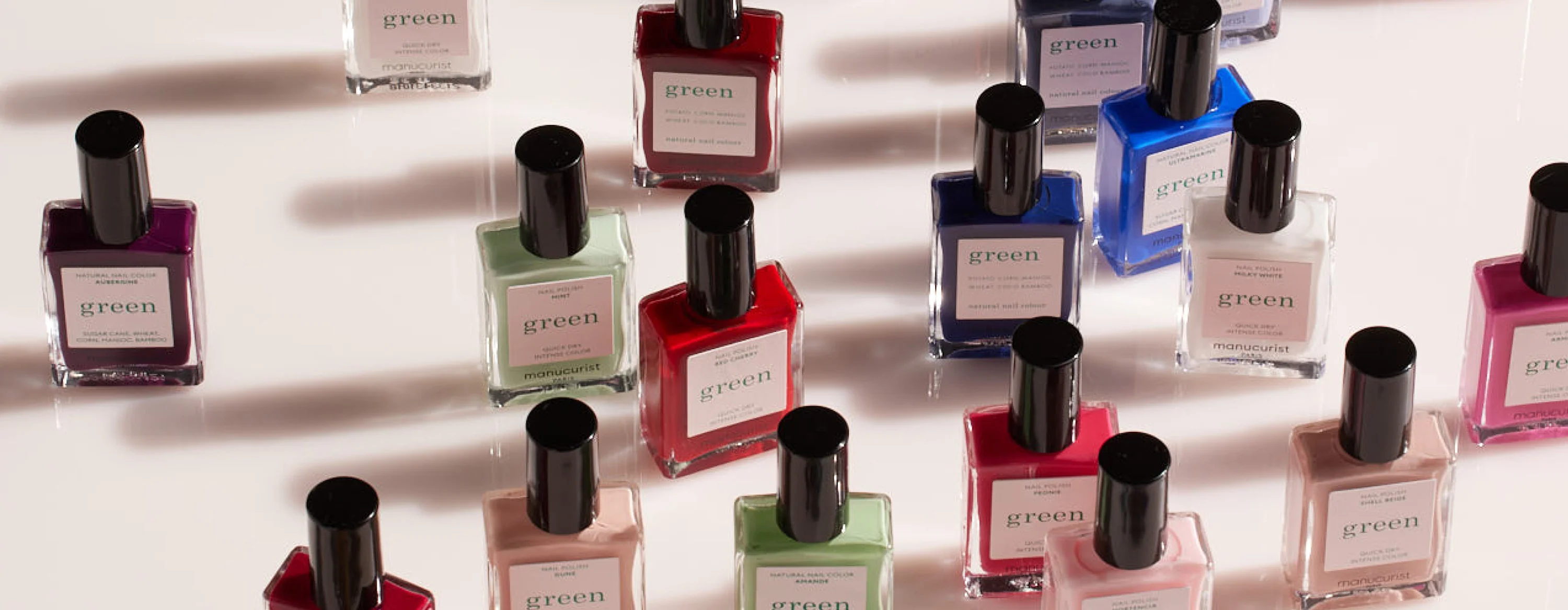 Bottles of nail polish in various colors, including red, blue, pink, green, and purple, are arranged in rows on a light surface. Each bottle has a black cap and a white label.
