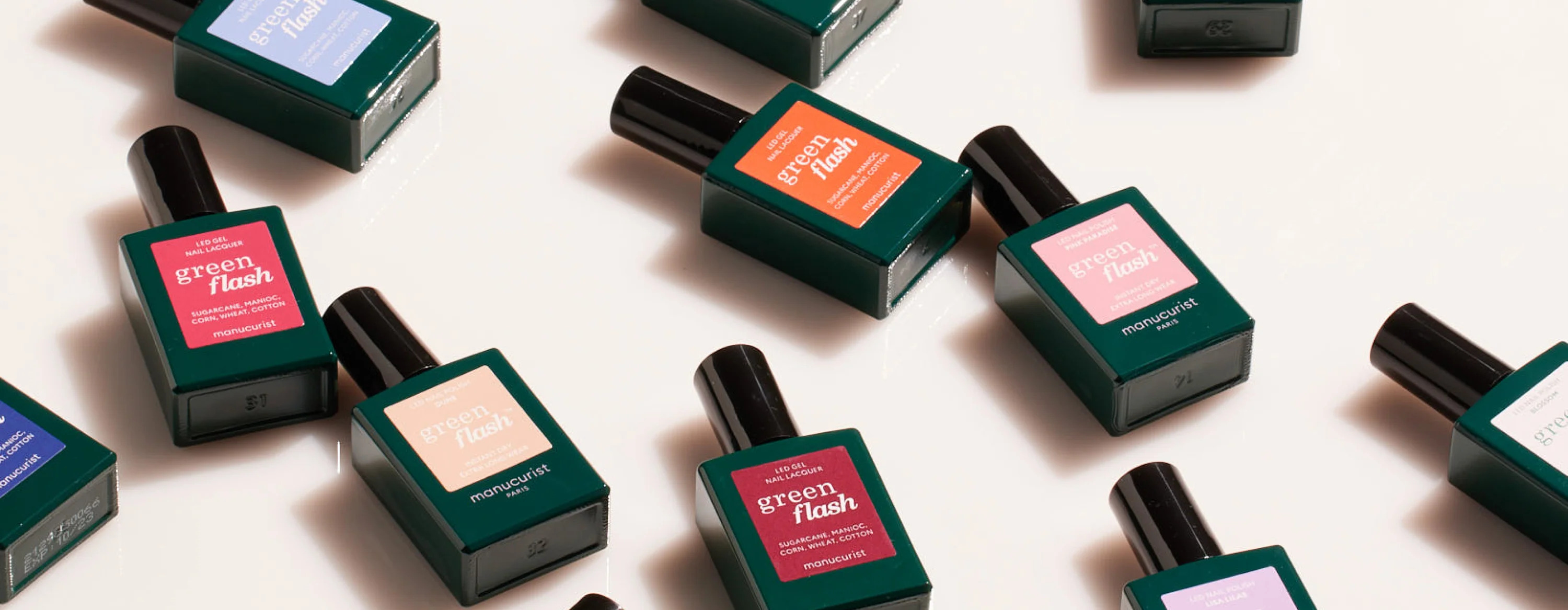 Multiple bottles of Manucurist Green Flash nail polish in dark green containers with various colored labels are arranged diagonally on a light surface.