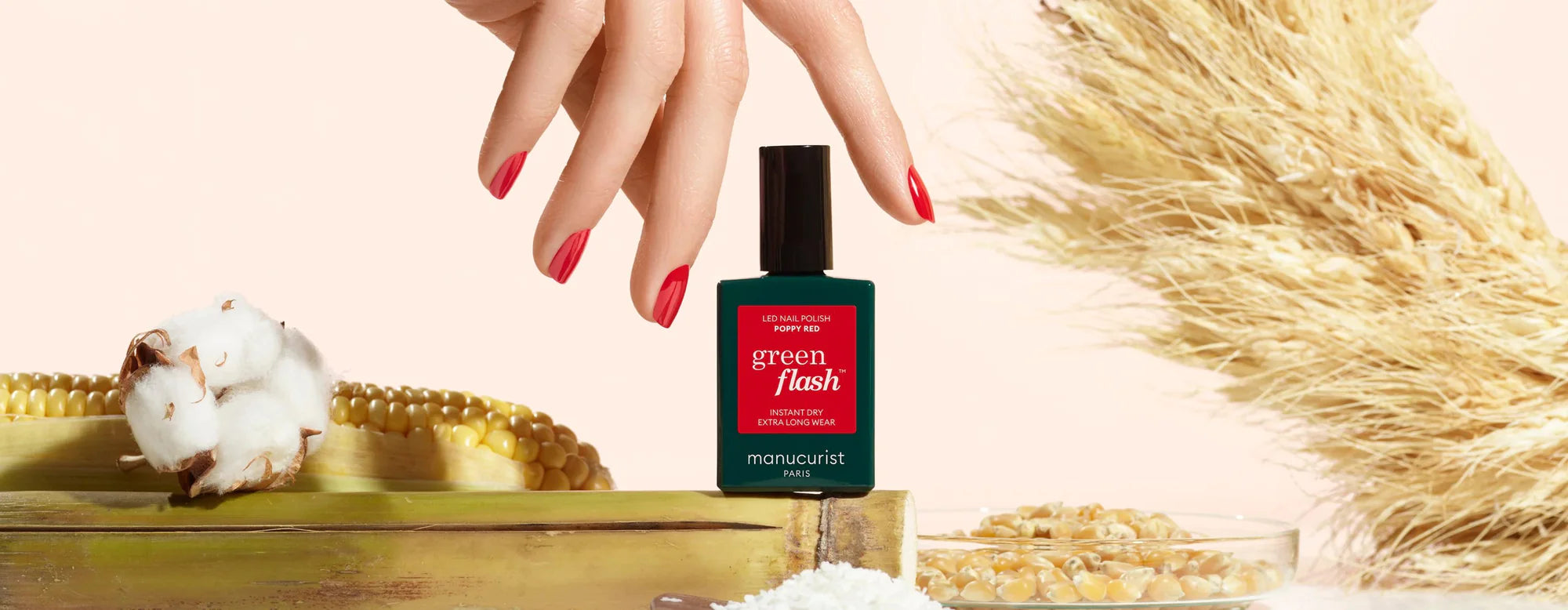 A hand with red nails holds a Green Flash nail polish bottle. Surrounding it are corn, cotton, grains, white petals, and dried plants, all on a light background.