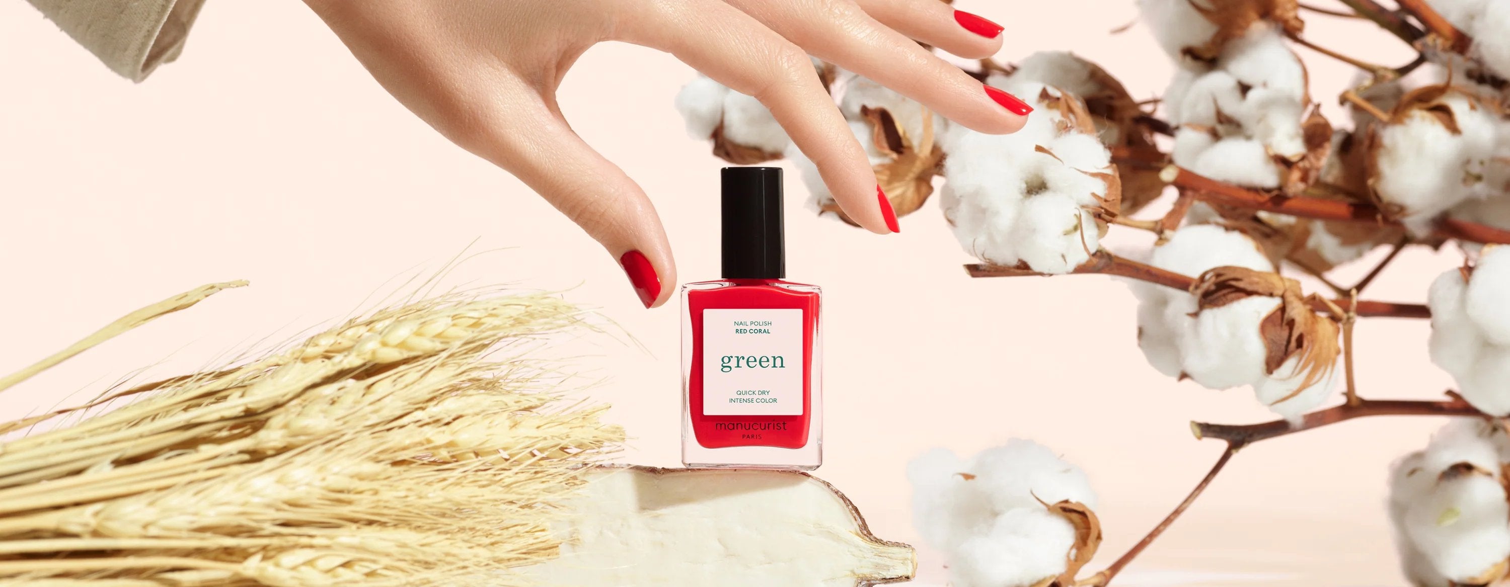 A hand with red-painted nails reaches for a red nail polish bottle labeled green placed among cotton branches and wheat stalks on a light background.