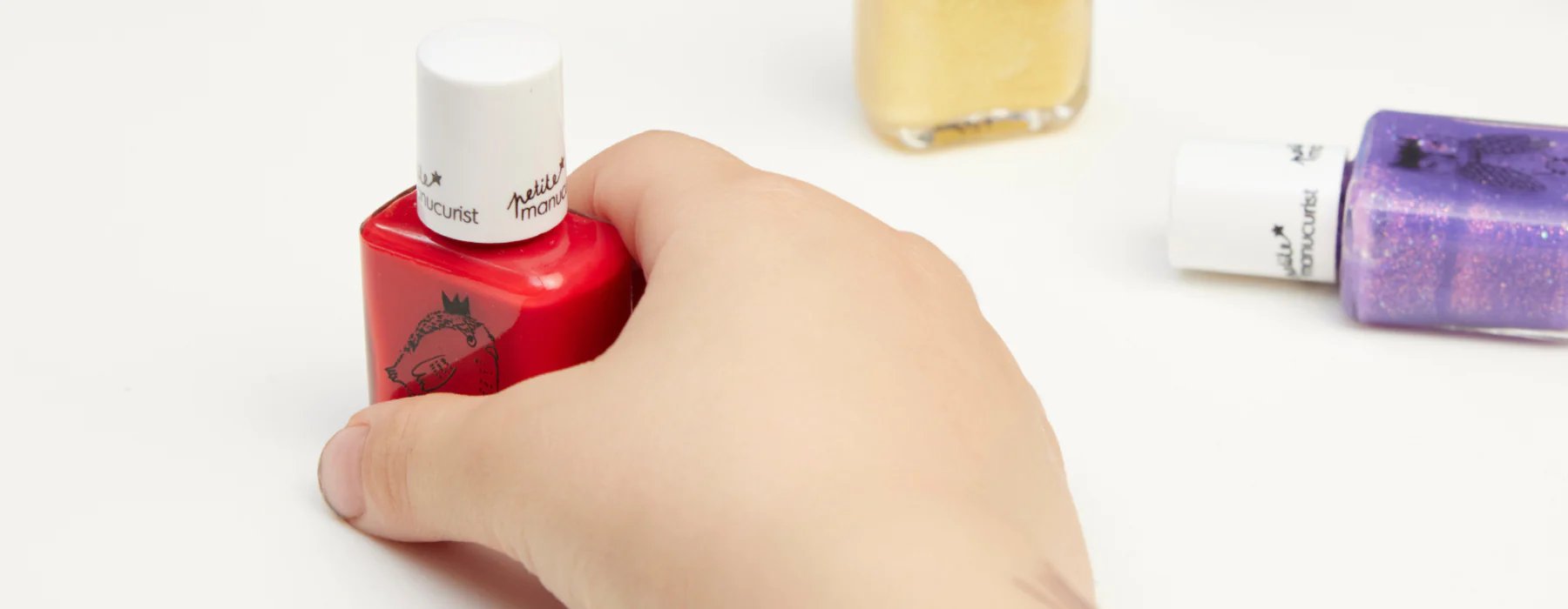 A childs hand holding a red nail polish bottle on a white surface, with yellow and purple nail polish bottles nearby.