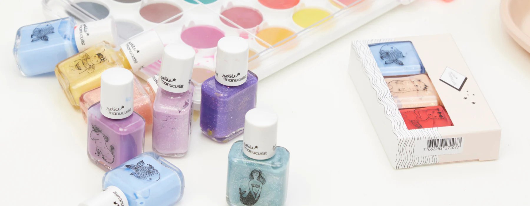 Several colorful nail polish bottles with illustrated labels are arranged near a watercolor paint palette and a boxed set of two nail polishes, all on a white surface.