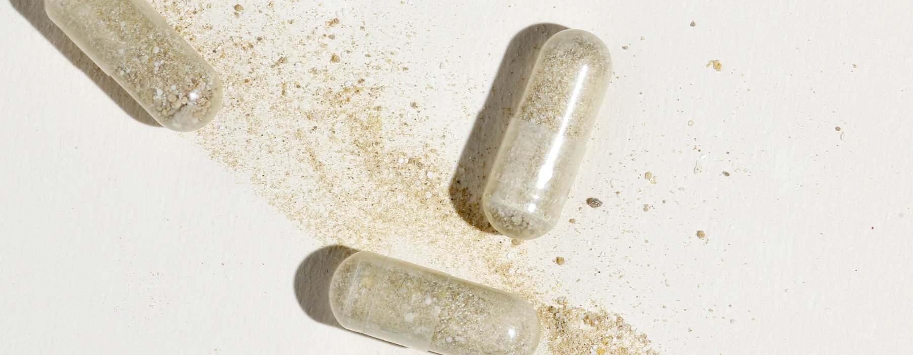 Three clear capsules filled with a beige, powdery substance rest on a white surface, with some of the powder spilled around and between the capsules.