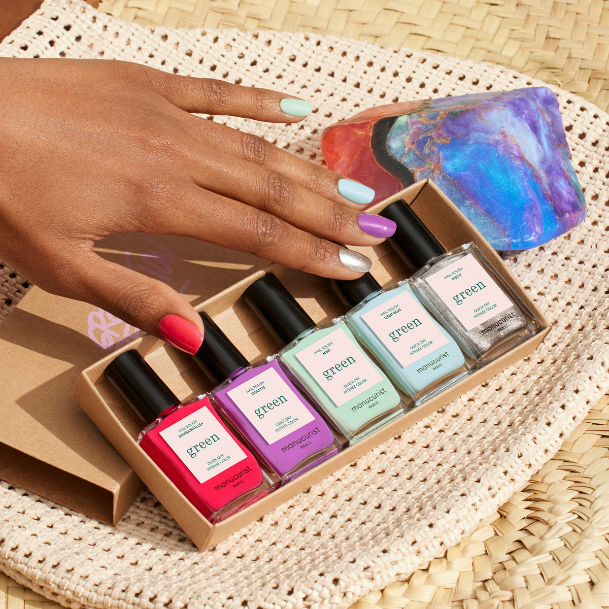 A hand with multicolored nails featuring Manucurist’s Retro Rainbow nail art holds an open box of five Green Flash bottles from the Summer Love collection, displayed on a woven mat beside a colorful rock. Each nail matches a polish shade in the set.
