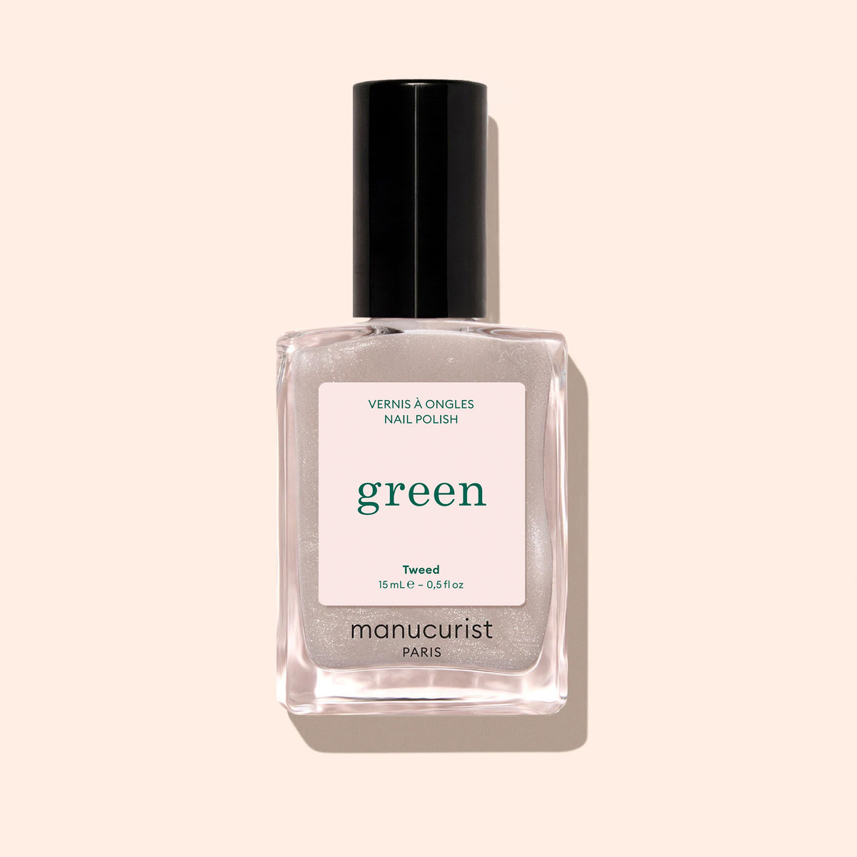 A bottle of Manucurist Tweed nail polish, from the Green line, features chic grey polish with a pearly sheen in a clear rectangular bottle with a black cap and white label, set against a light beige background.