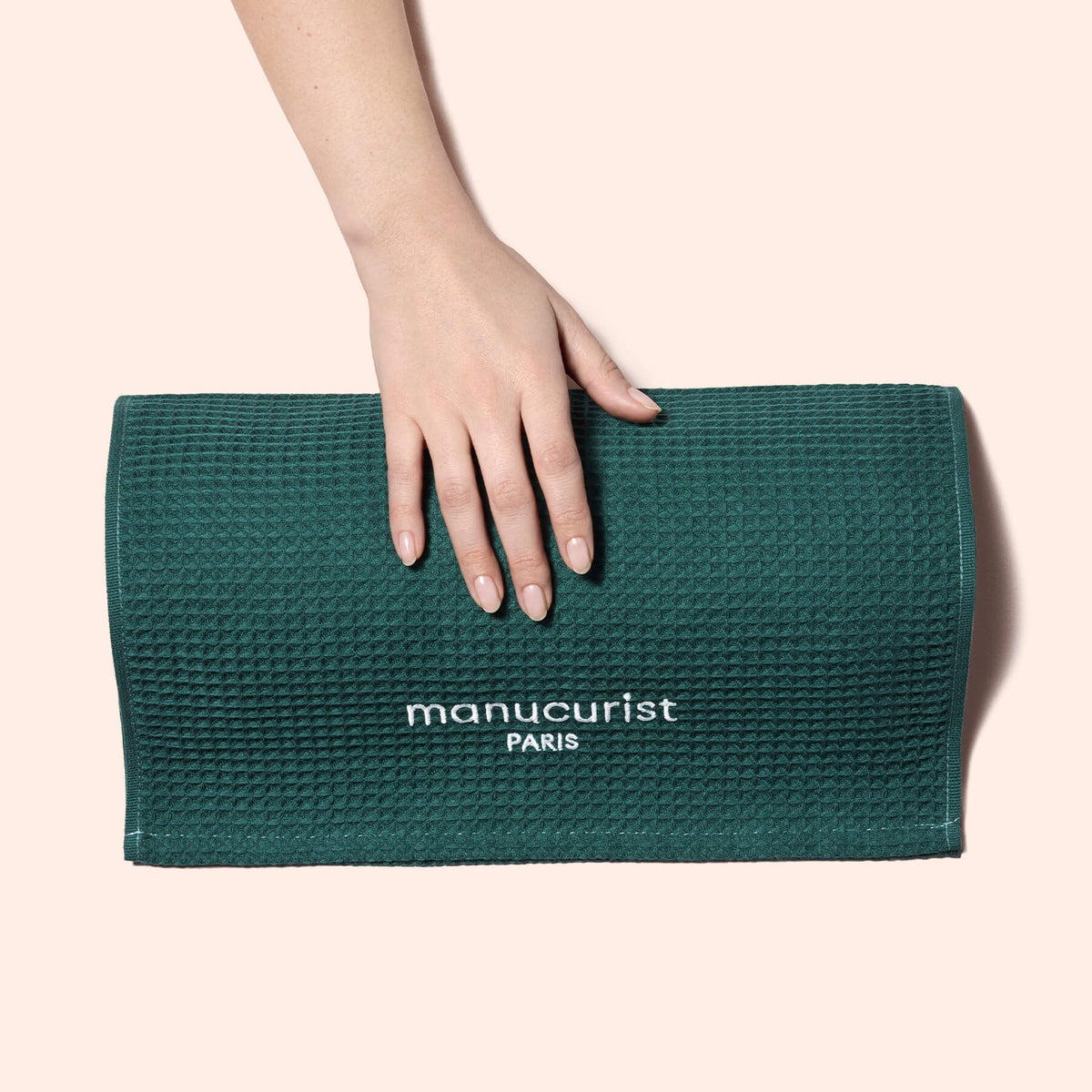 A manicured hand rests on a textured dark green Manucurist Towel, made from organic cotton and embroidered with "manucurist PARIS" in white, shown against a light beige background.