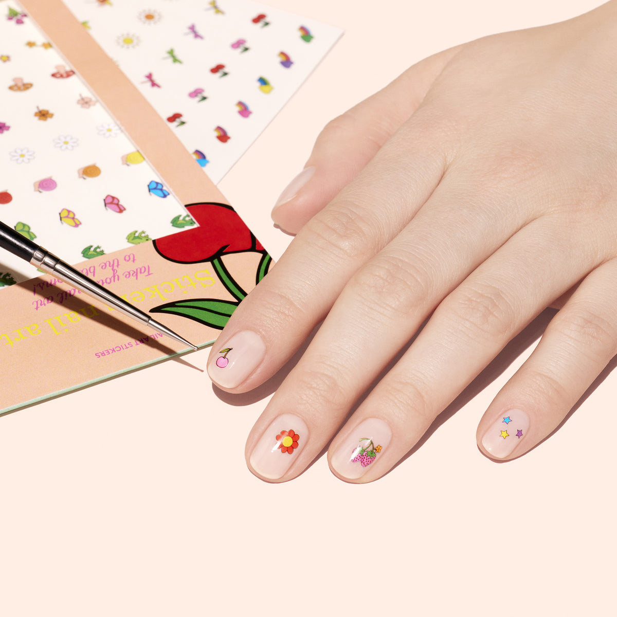 A hand shows off nude polish and Manucurist Nail Art Stickers with colorful florals. Nearby, nature-themed sticker sheets and a thin brush rest on a peach backdrop—perfect chic summer nail inspiration.