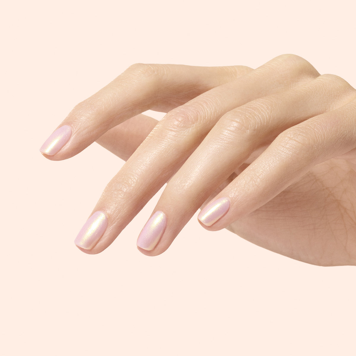 A hand with neatly manicured nails painted in Manucurist’s Golden Glazed Powder, a light iridescent pink polish with pearly gold shimmer, is shown against a pale solid background.