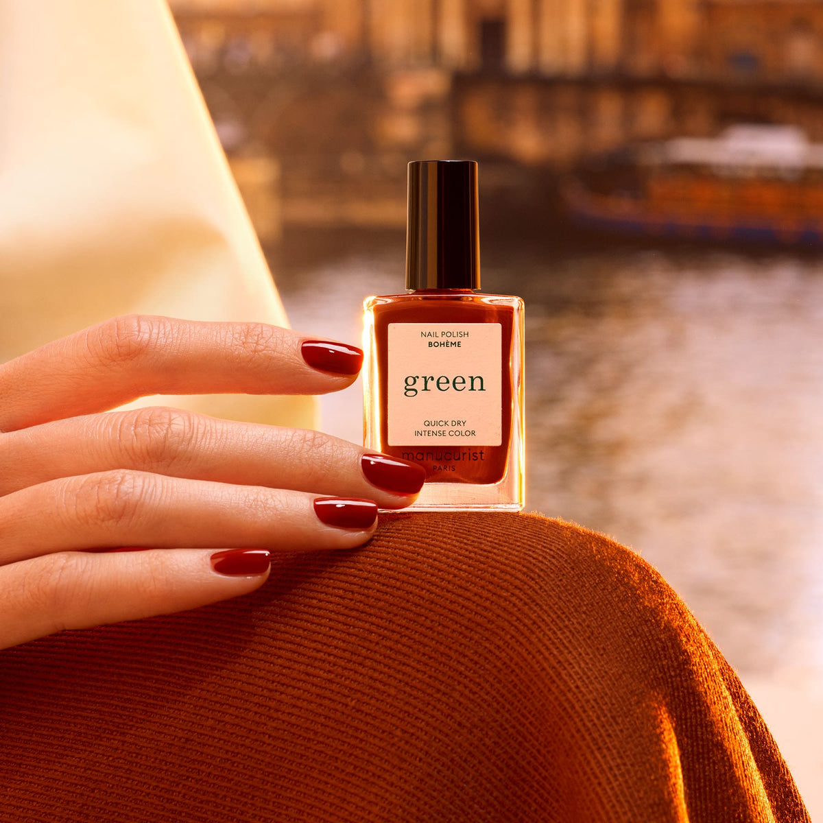 A hand with dark red polished nails holds a bottle of Manucurist Bohème polish outdoors, bathed in golden light with blurred water and buildings in the background.
