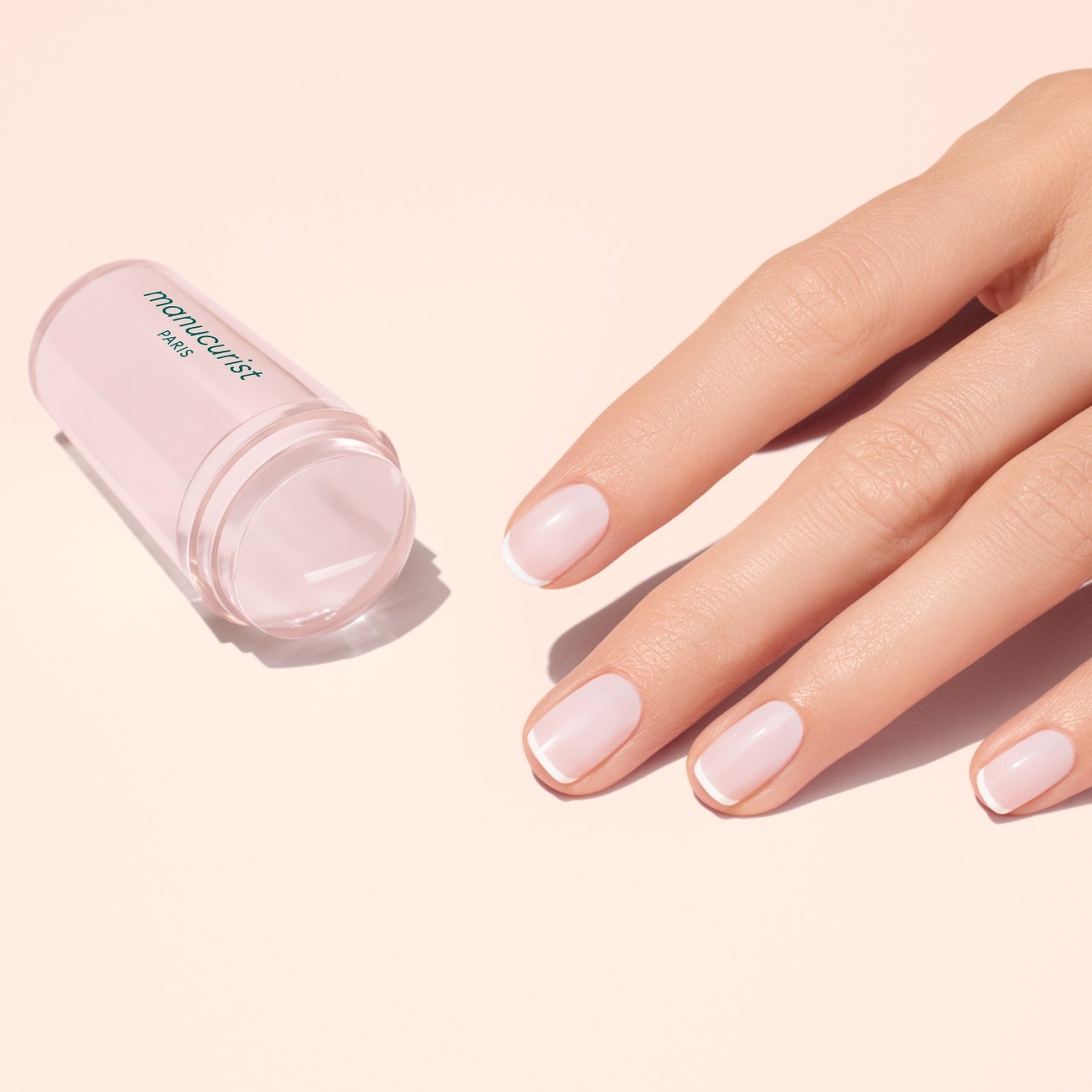 A hand with pale pink, glossy nails rests beside a translucent pink Mani stamper. The French Active™ Kit is perfect for creating flawless French tips at home.