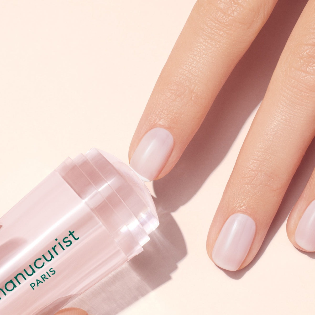 A hand with neatly manicured nails uses the Manucurist Mani Stamper, a clear cylindrical nail tool, perfect for flawless French tips, set against a light pastel background.