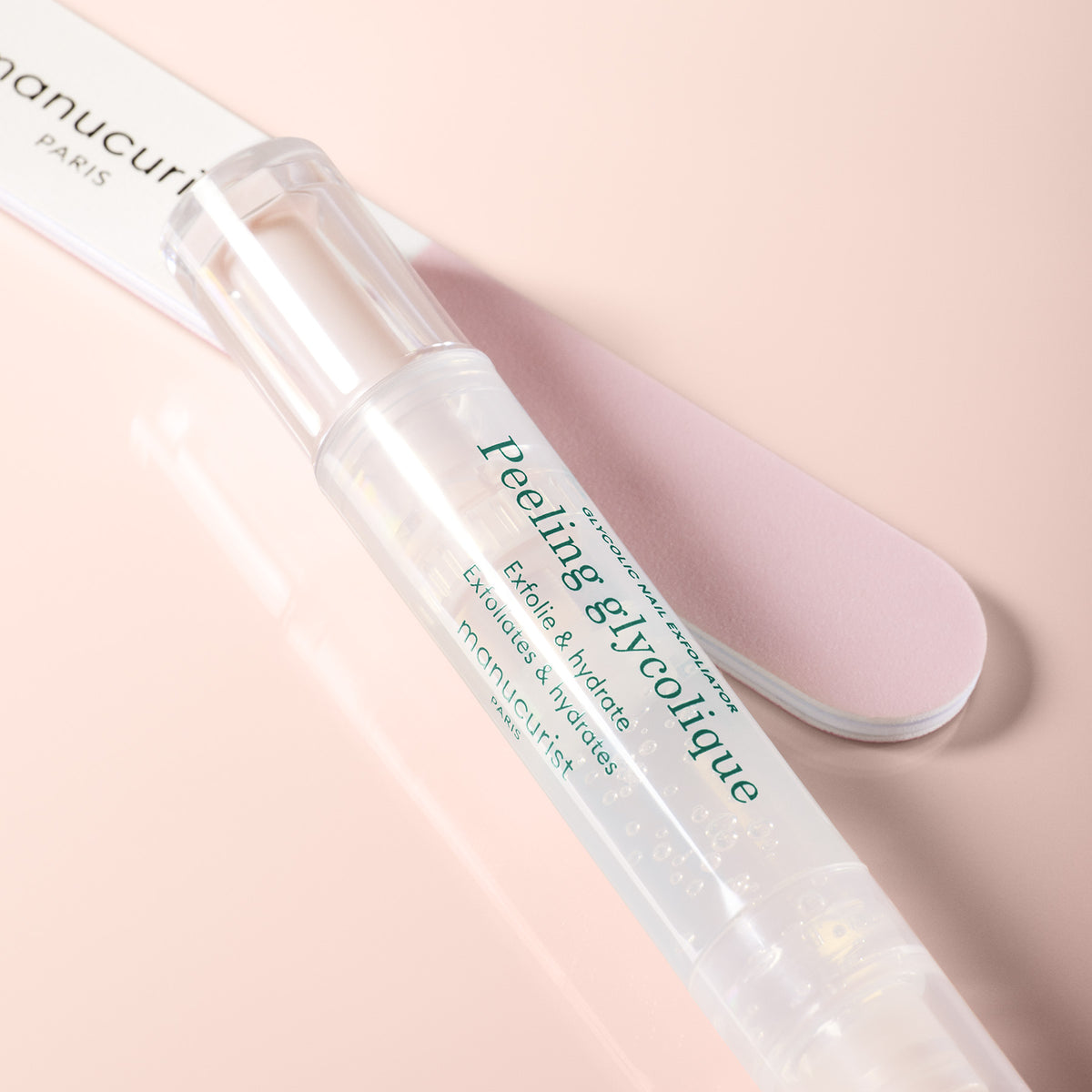 A clear tube labeled Glossy Booster Duo with a white cap rests on a light pink surface next to a pink and white nail file. This Manucurist set is perfect for exfoliating and hydrating, leaving your nails glossy.