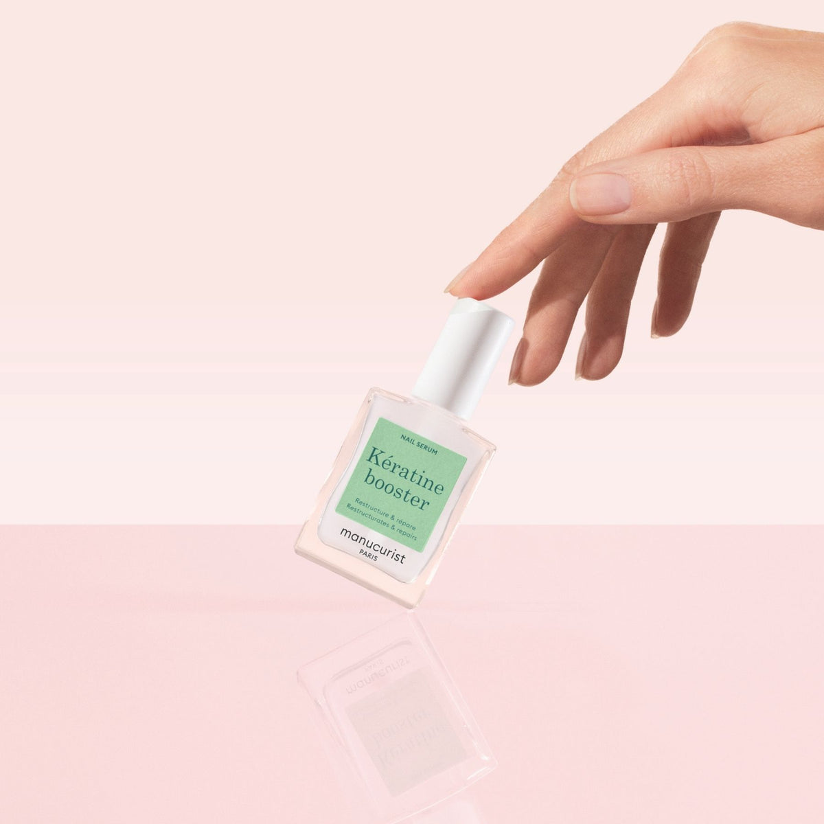 A manicured hand tips a bottle of Manucurist Nail Strengthening Serum on a glossy pink surface, set against a light pink background—ideal for enhancing nail care and restoring damaged nails.