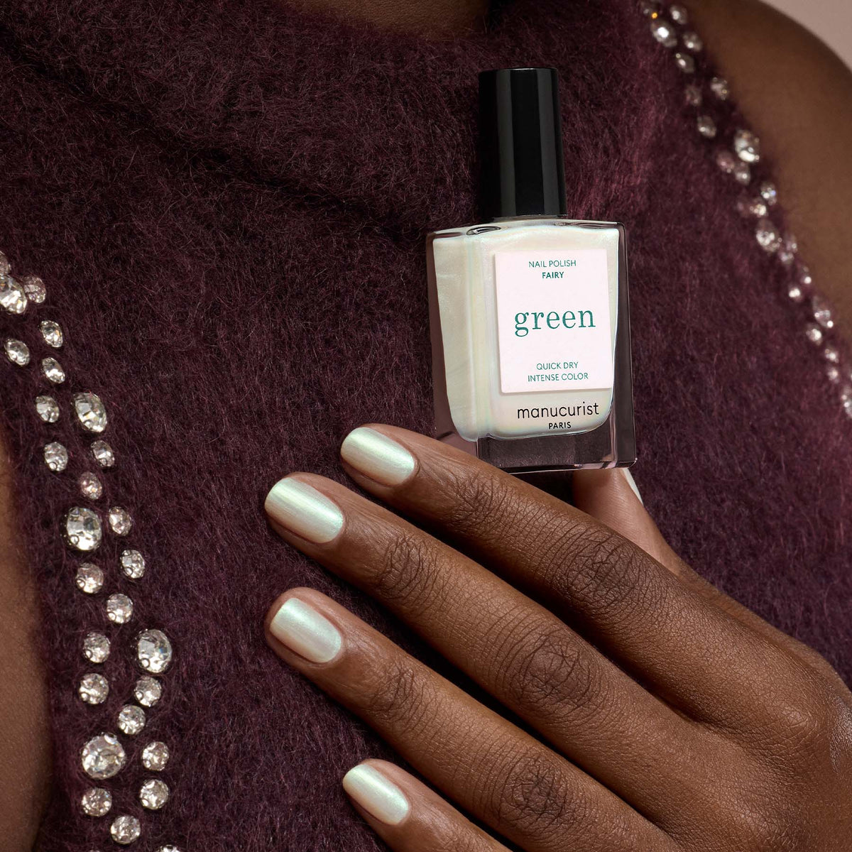 A person with light green painted nails holds a bottle of Manucurist “Fairy” nail polish against their maroon, bejeweled sweater.