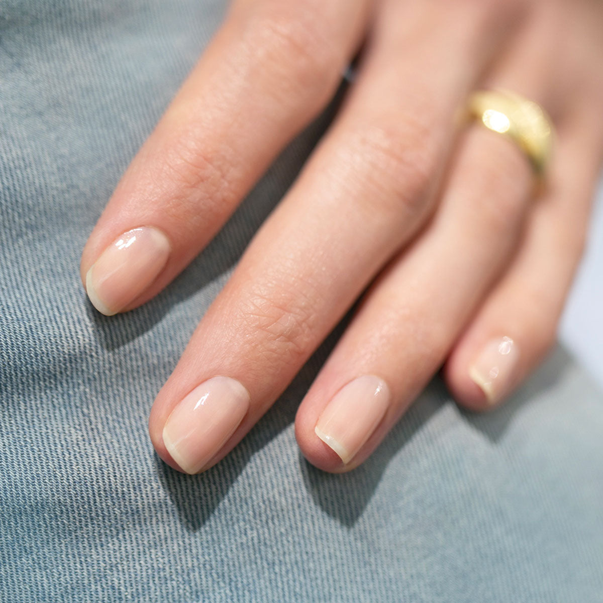 A close-up of a hand with short, natural nails painted with Manucurist’s Green™ nail polish in Beige. The glossy nude shade shines against light blue denim and is complemented by a gold ring on one finger.