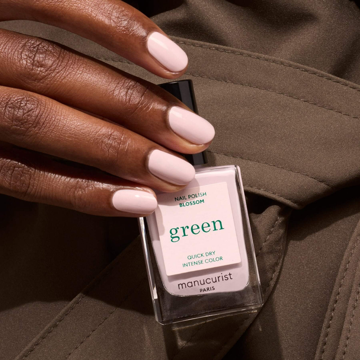 A hand with neatly manicured, light pink nails holds a bottle of Manucurist Blossom nail polish against an olive-green fabric background.