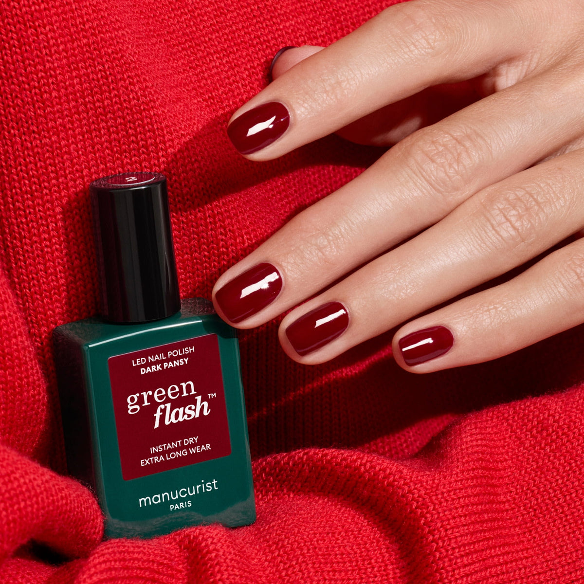 A hand with short, glossy, dark red nails rests on red fabric beside a bottle of Manucurist Dark Pansy gel polish.