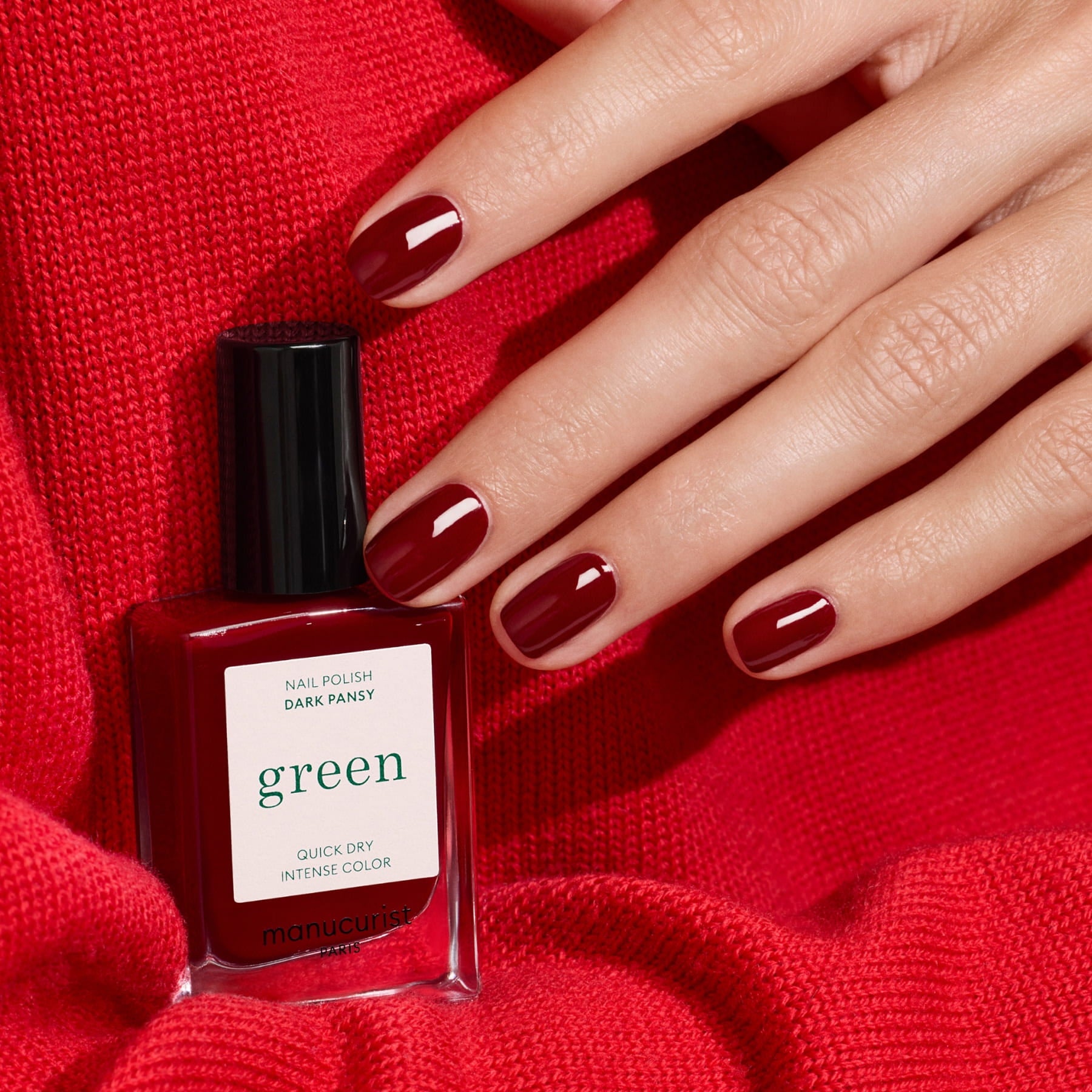 A hand with short, manicured nails painted in Manucurist’s Dark Pansy polish rests on red fabric, next to a bottle of Manucurist Dark Pansy from the Green range in a matching shade.