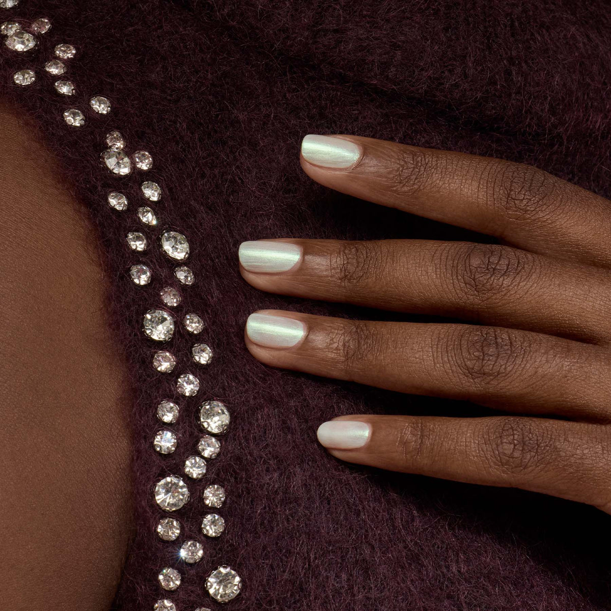 A hand with Manucurist’s Fairy pearly white gel polish rests on burgundy fabric adorned with sparkling rhinestones arranged in a curved line.