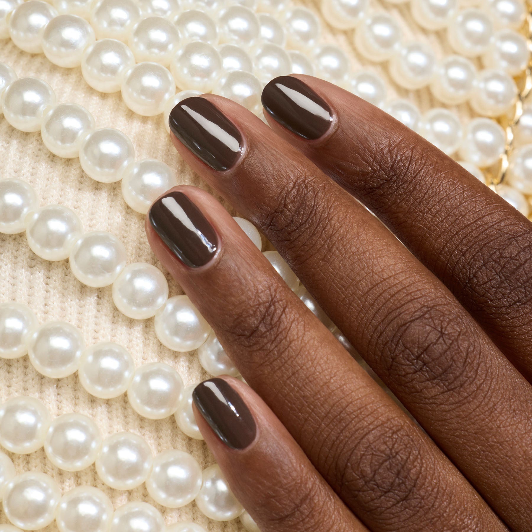 A hand with neatly manicured Mocha gel polish nails by Manucurist, in a cool dark brown shade, rests on a surface adorned with white pearls and cream fabric.