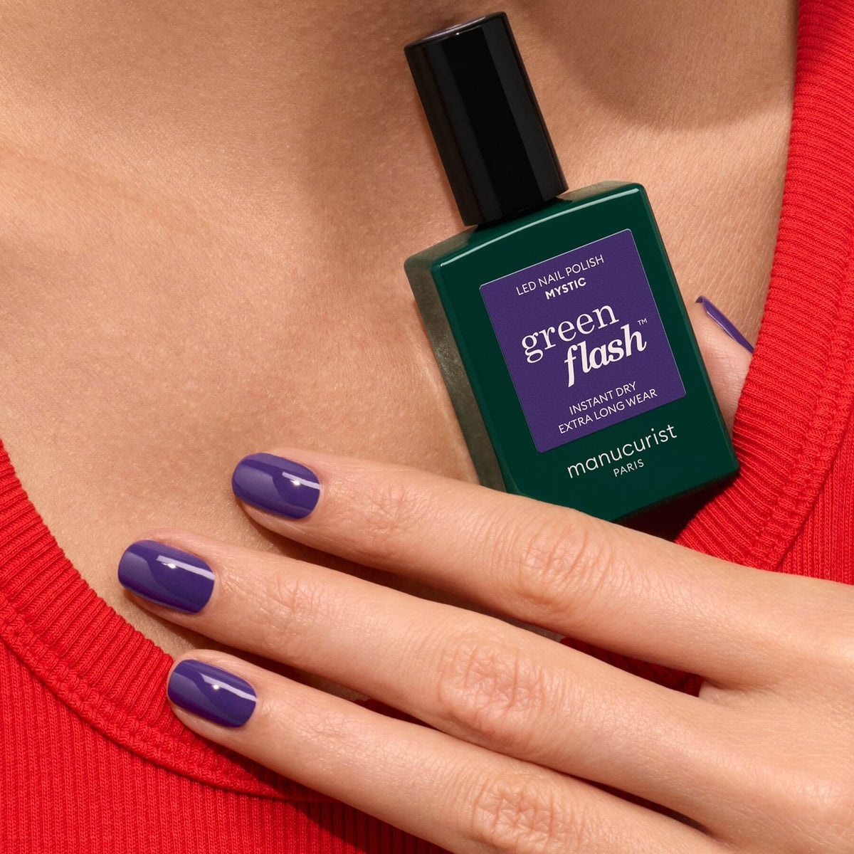 A hand with neatly manicured nails painted in Manucurist’s Mystic holds a bottle of the same polish, set against a rich red fabric background.