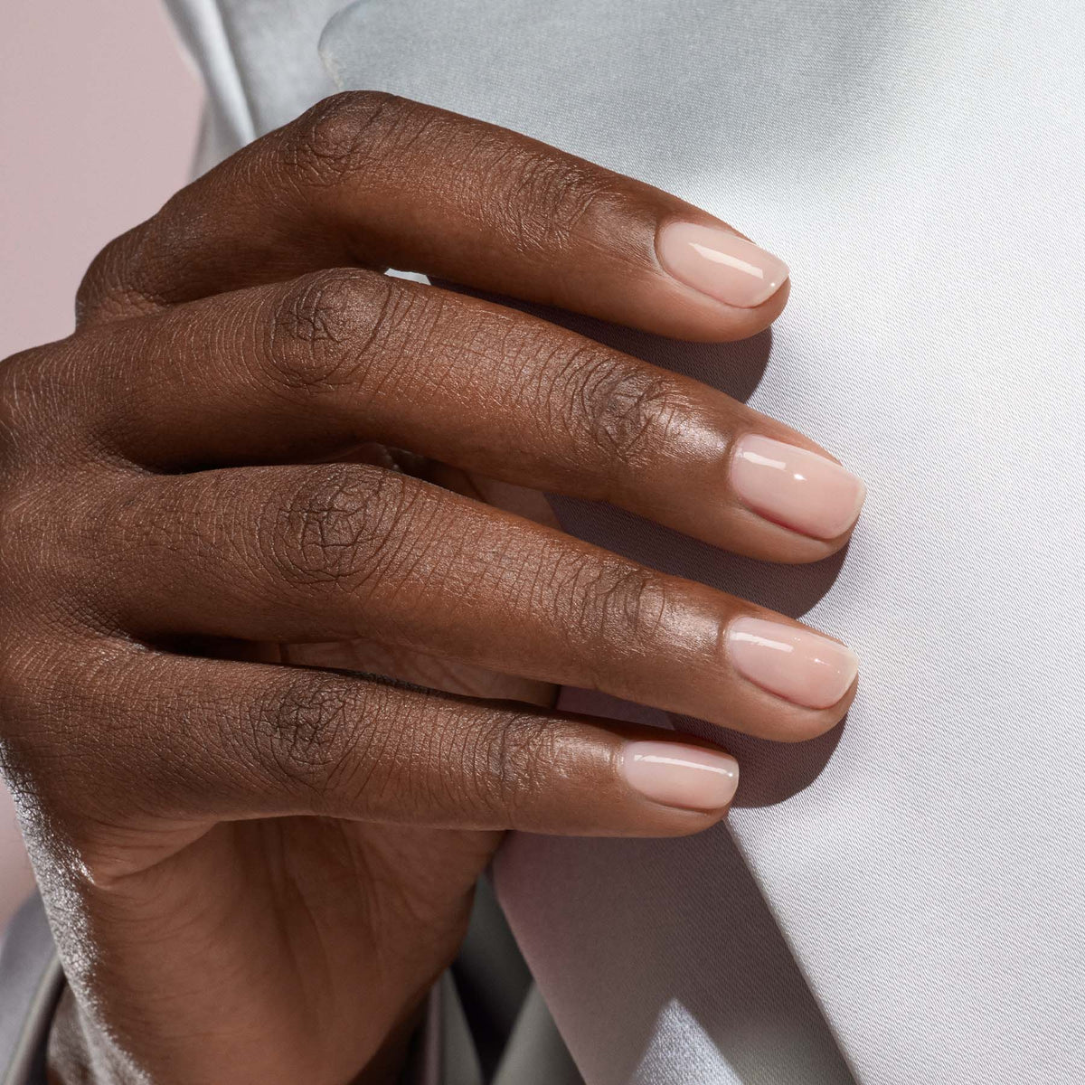 A close-up of a hand with neatly manicured nails painted in Manucurist-test’s Pale Rose - Free sheer pastel pink nail polish, resting gently on light-colored fabric.