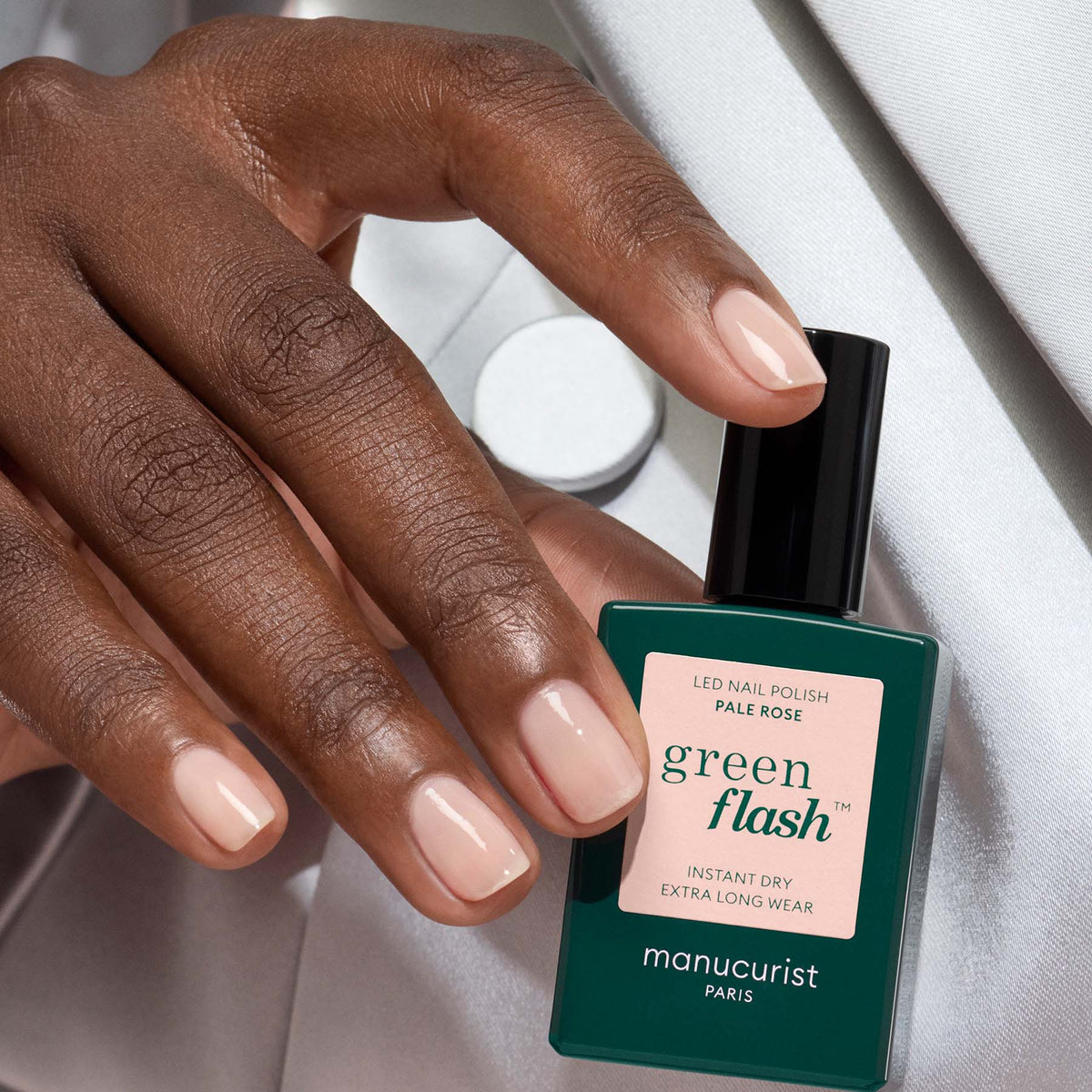 A hand with neatly manicured, soft sheer rose nails holds a bottle of Manucurist Pale Rose polish, set against a light gray satin background.