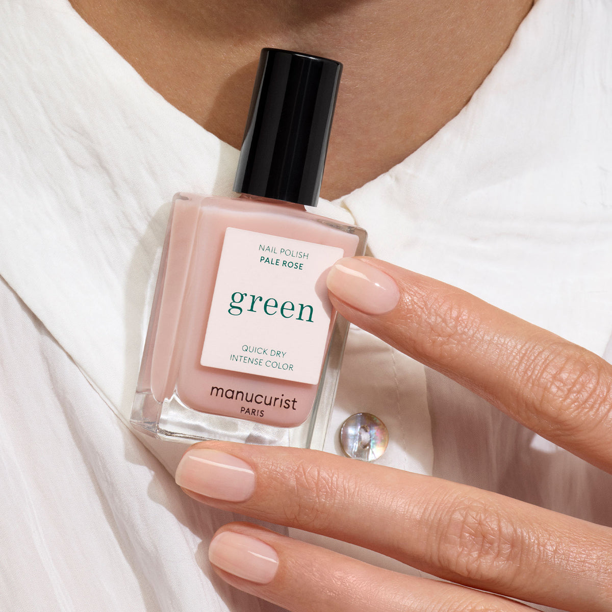 A hand with neatly manicured, sheer pastel pink nails holds a bottle of Manucurist Pale Rose nail polish against a white shirt.