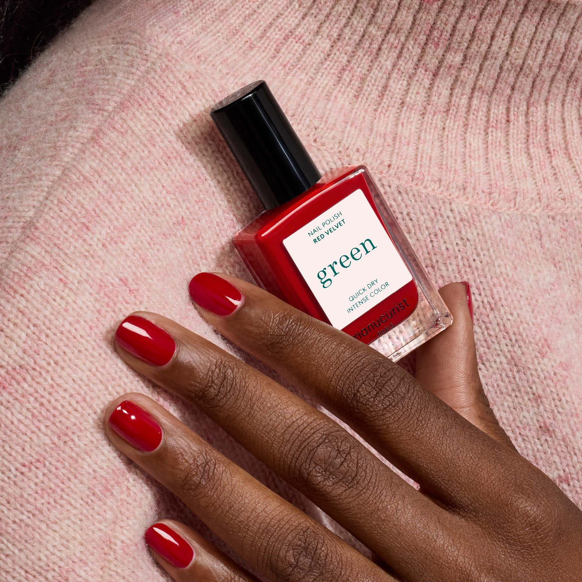 A hand with neatly manicured nails in Red Velvet by Manucurist holds the bottle against a soft, pink knitted fabric background.