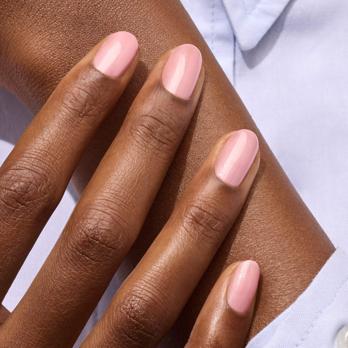 A close-up of a hand with neatly manicured nails painted in Satin by Manucurist, a pale pink LED gel polish. The medium brown skin contrasts with a light blue shirt, highlighting the elegant semi-permanent manicure.