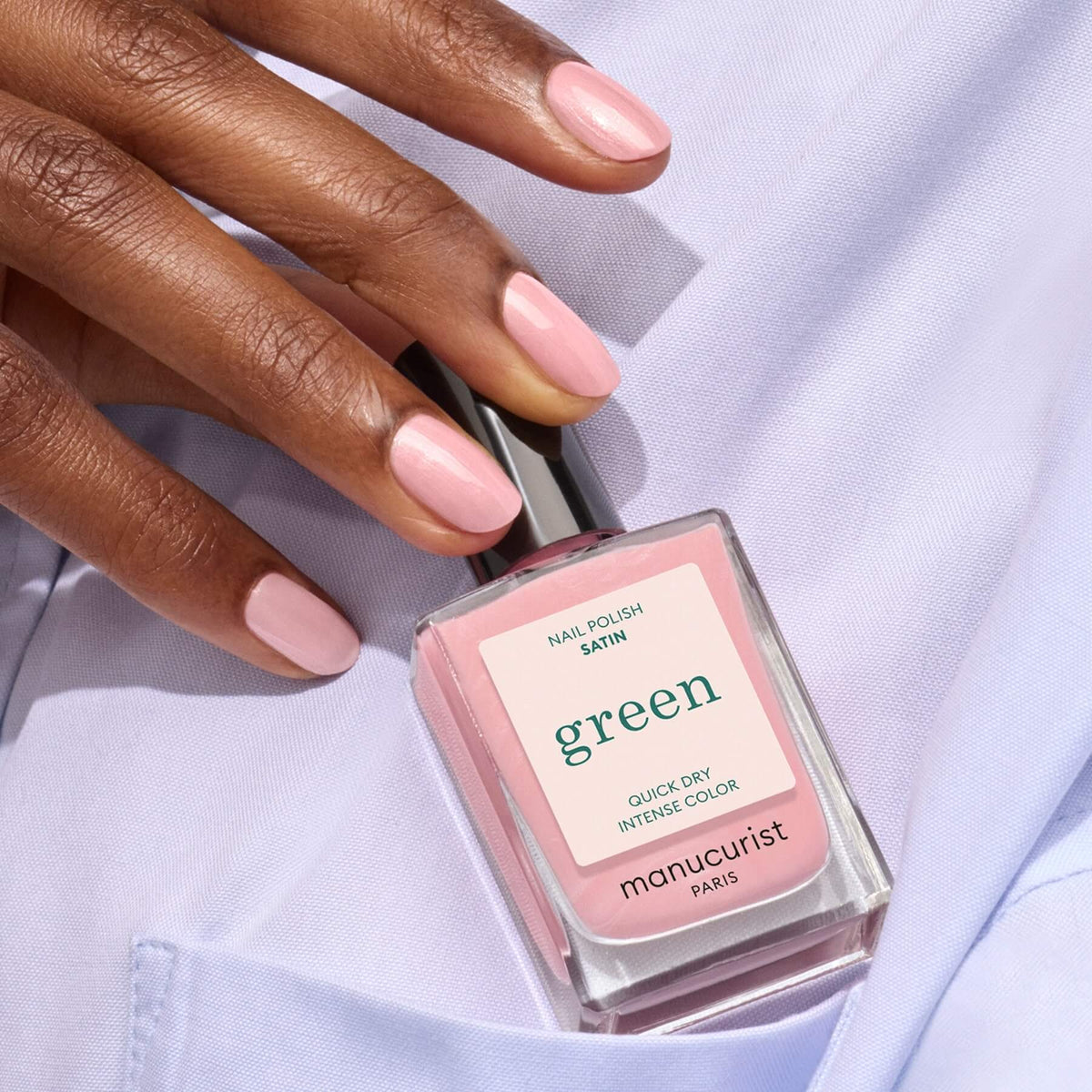 A hand with neatly manicured nails painted pale pink holds a bottle of Manucurist Satin nail polish in a matching shade, resting on light lavender fabric.
