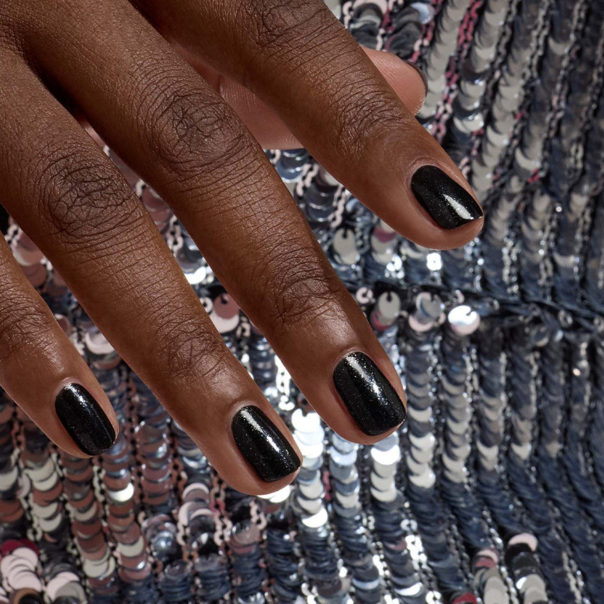 A hand with neatly manicured nails painted in Manucurist’s Sparks, a sparkling black polish, rests on a surface covered in shiny silver sequins.