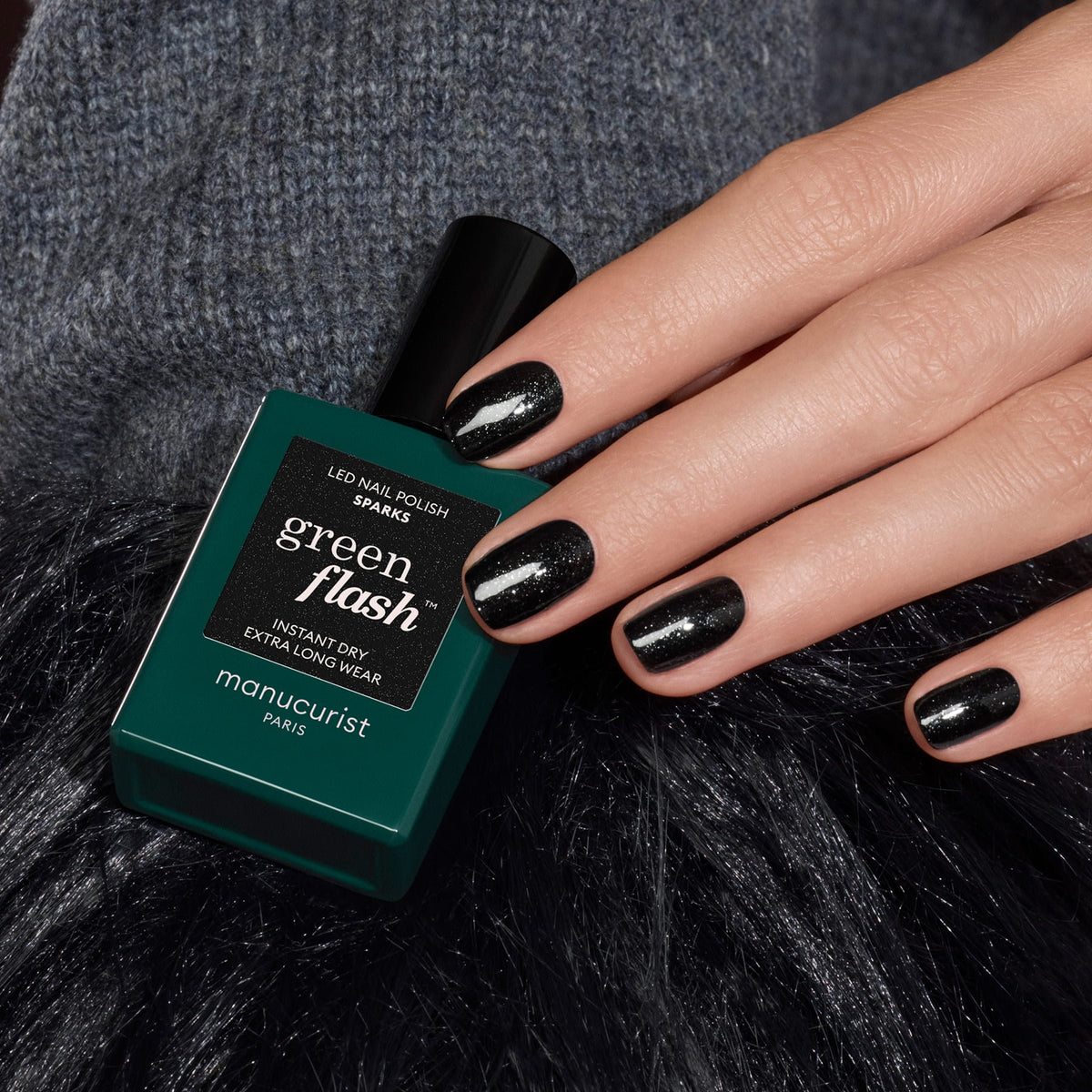 A hand with short, oval nails painted in shimmering black gel polish holds a Manucurist Green Flash nail polish bottle labeled "Sparks" against a textured dark background.