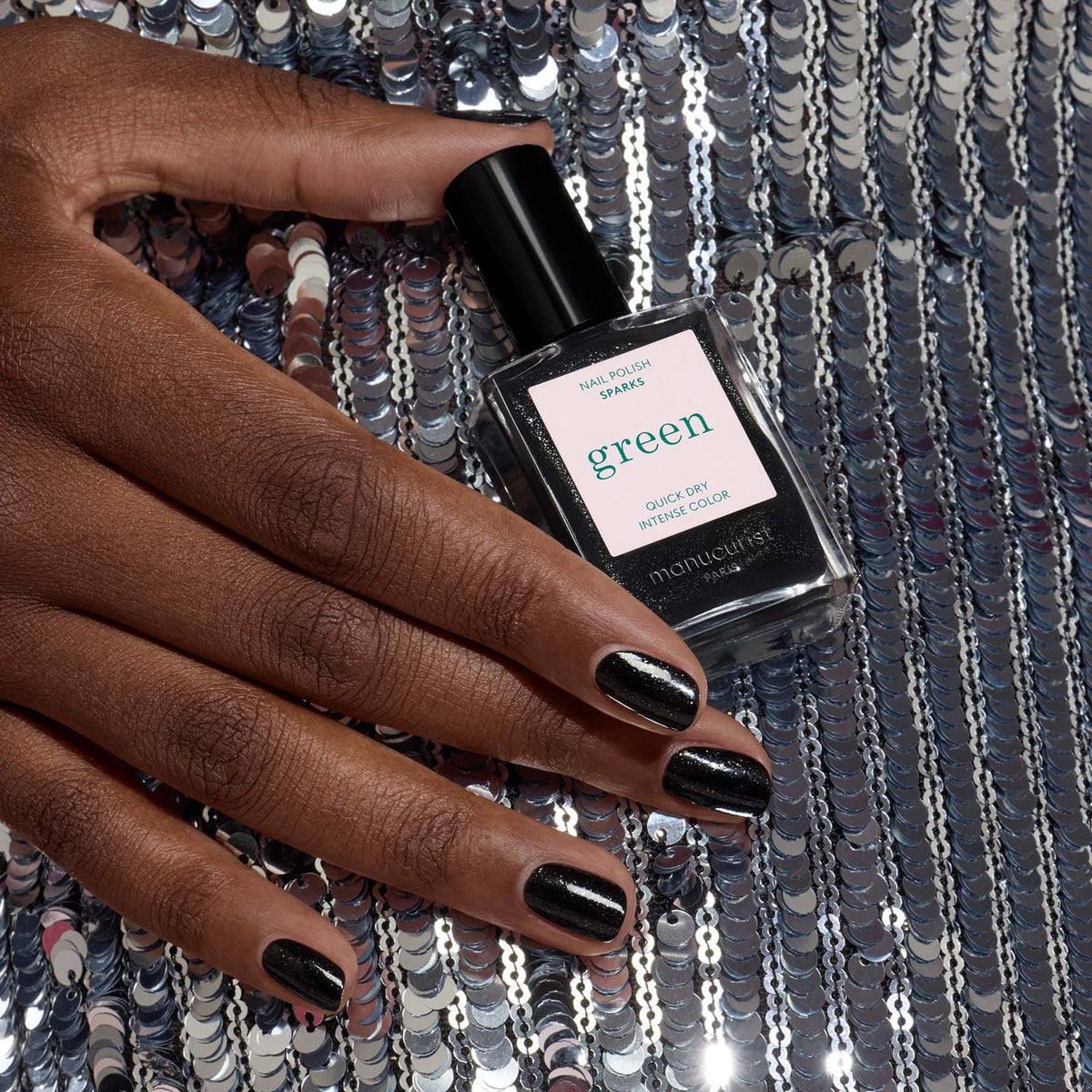 A hand with sparkling black nail polish named Sparks by Manucurist rests on silver sequin fabric, holding the bottle.