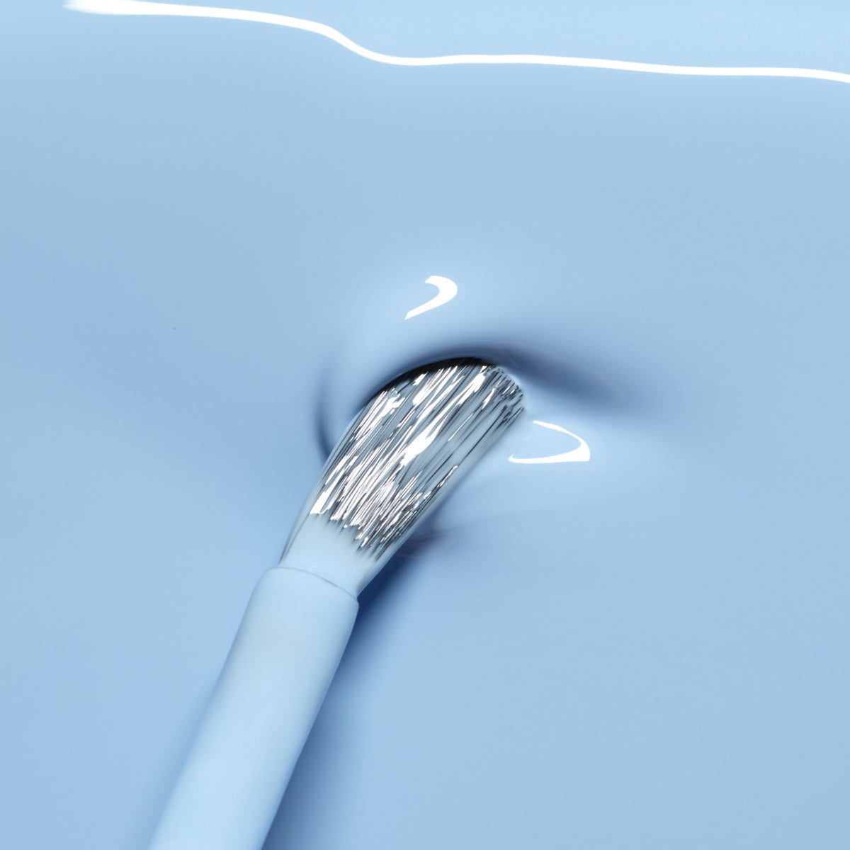 A close-up of the Manucurist Bubble brush dipped into glossy light blue nail polish with grey undertones, as ripples and reflections shimmer on the liquid surface.