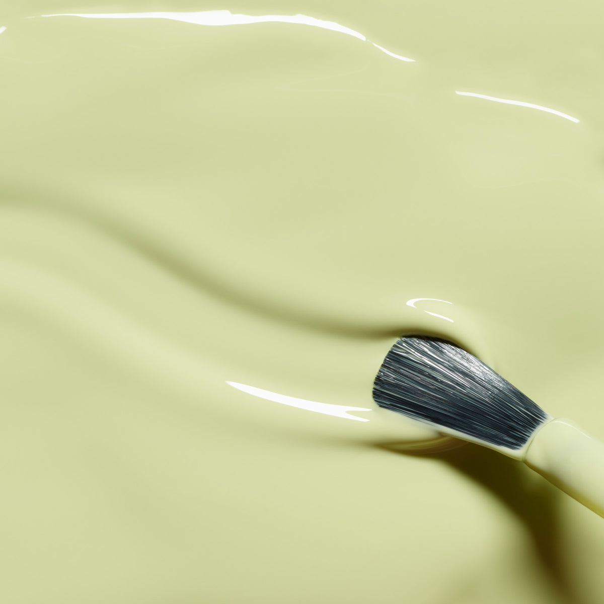 A close-up of a makeup brush partially submerged in Manucurist Pistachio foundation, showcasing its smooth, creamy light yellow shade with yellow undertones and a gently rippled texture.