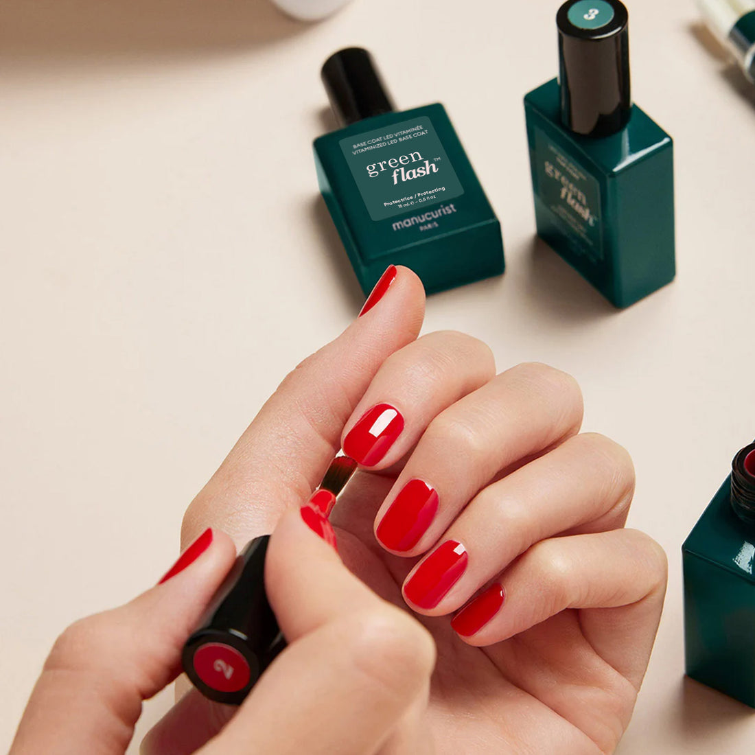 A person applies bright red nail polish, finishing with Manucurist Trio Base Top Remover Green Flash™ for a glossy look. Several green bottles of Manucurist polish are displayed on a beige surface in the background.