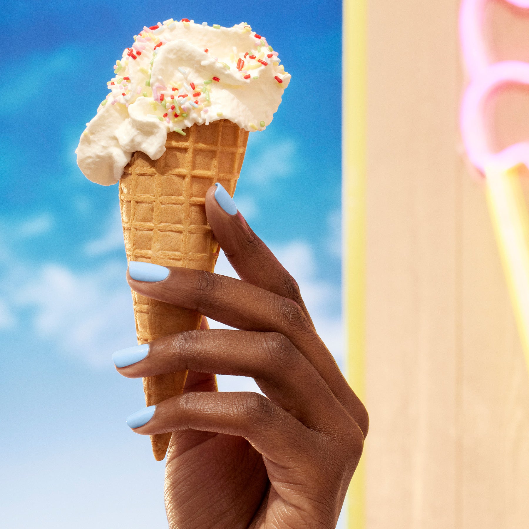 A hand with Bubble by Manucurist, a light blue-grey nail polish, holds a waffle cone topped with vanilla ice cream, sprinkles, and whipped cream against a sunny sky and blurred neon sign.
