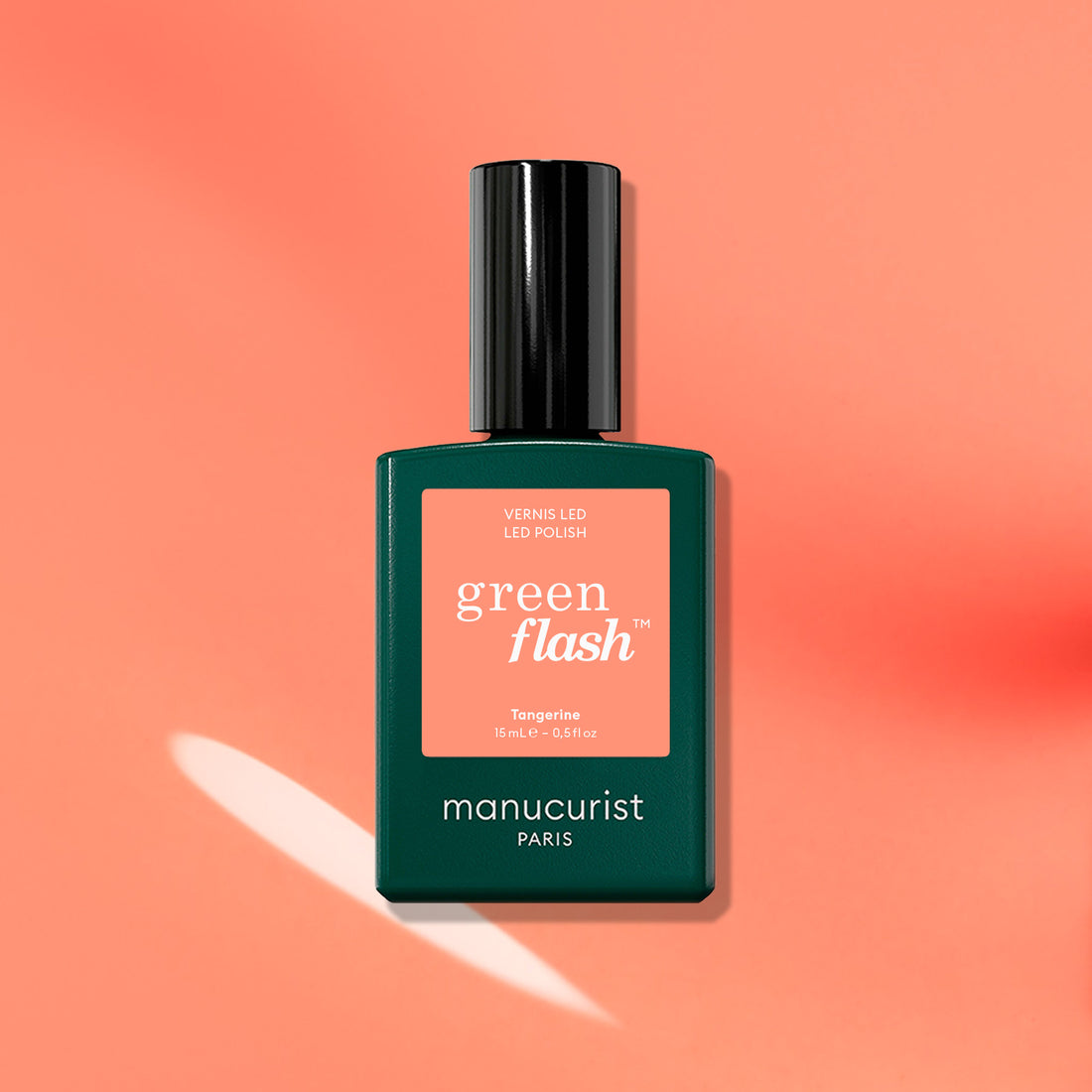 A Manucurist Tangerine gel polish bottle rests on a peach-orange background with white highlight and shadow. Its dark green bottle and black cap evoke ’90s vibes, while the neon orange shade pops boldly.