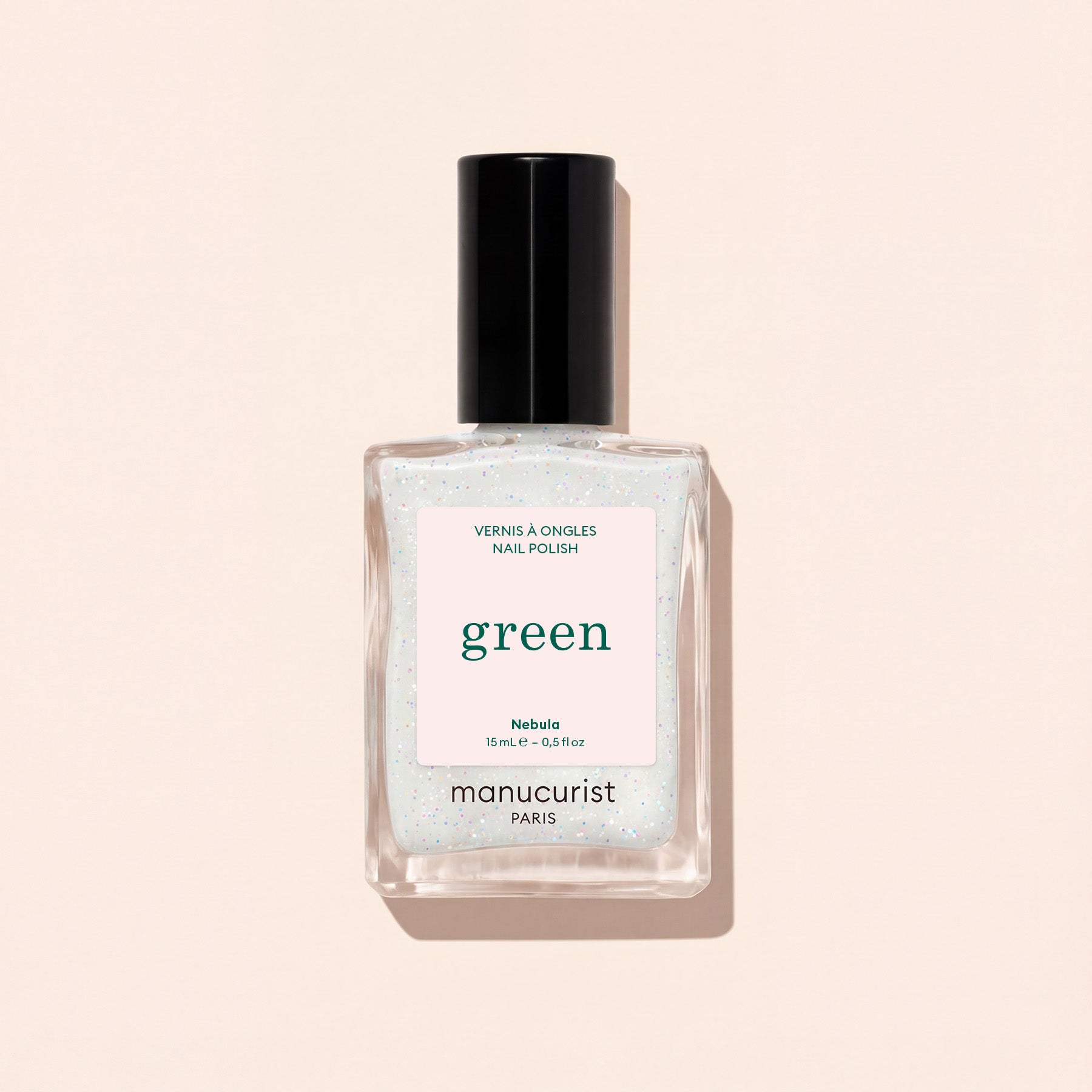 A clear glass bottle of Manucurist nail polish in the shade Nebula, featuring a black cap and white label, is displayed on a pale beige background.