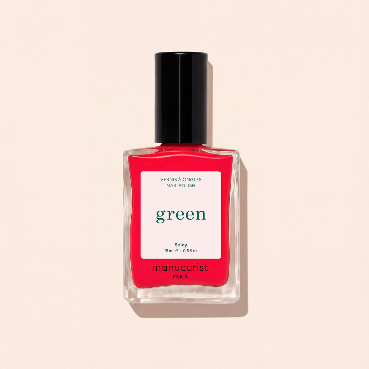 A clear glass bottle of Manucurist nail polish in the Spicy shade, featuring a black cap and “green” labeling, rests on a light beige background.