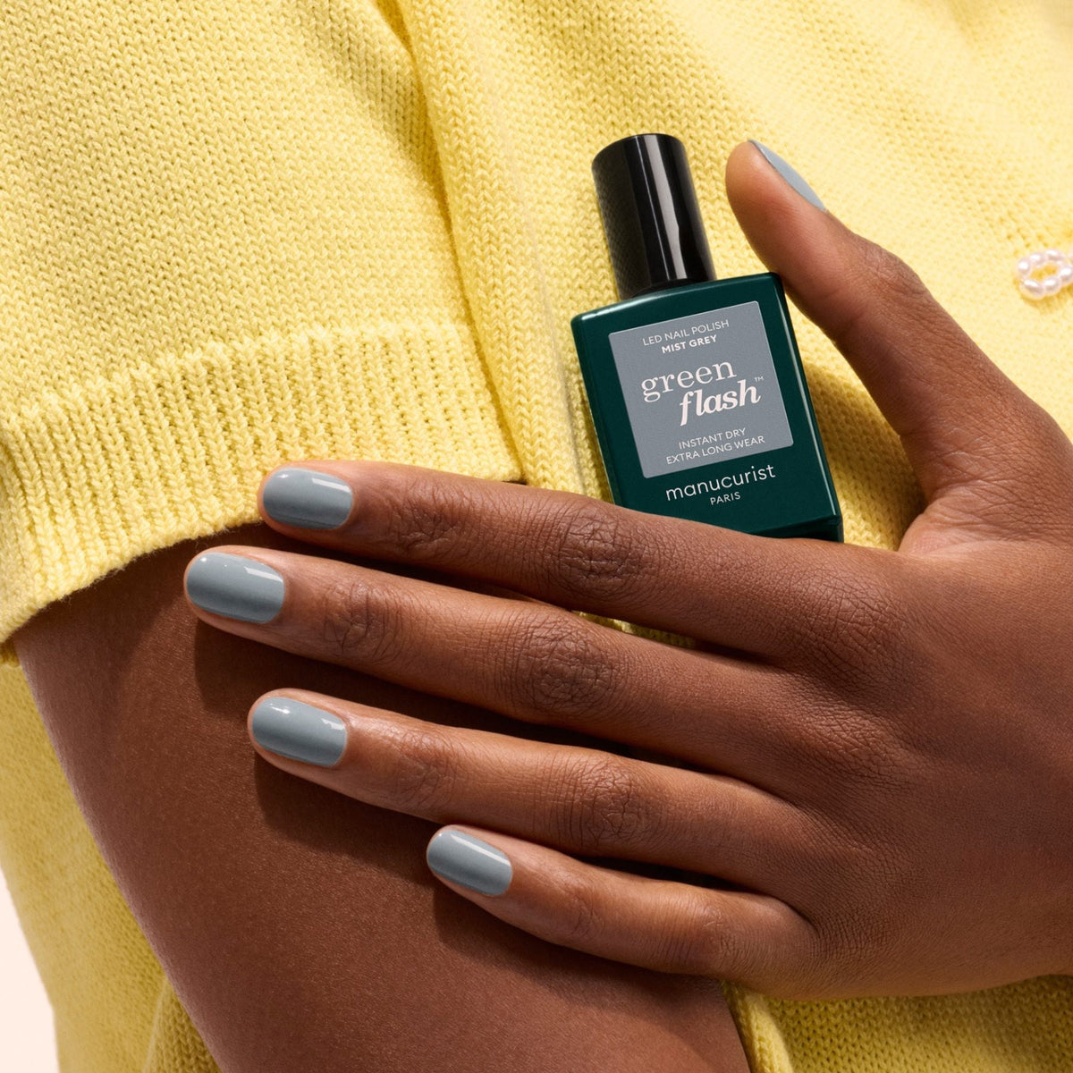 A person with medium-brown skin and light blue-gray, Mist Grey-painted nails holds a bottle of Manucurist gel polish against their yellow short-sleeve top.